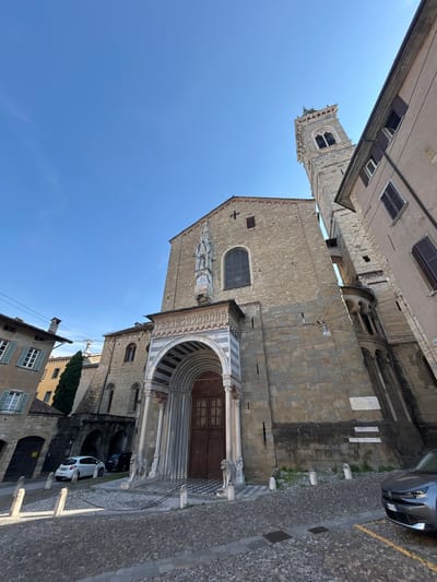 The image depicts a low-angle view of a stone church with a prominent bell tower, set against a clear blue sky. The church features an arched entryway with intricate carvings and marble columns. Two stone lion statues flank the entrance. The cobblestone street in front of the church has a checkered pattern leading up to the steps. A white car and a dark car are parked on the street. The architecture suggests a historical European city, likely Italy. The scene captures a sunny day with shadows indicating mid-morning or late afternoon light. The overall impression is one of historical significance and architectural grandeur.
