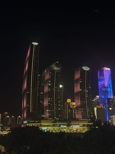 An image taken at night captures a cluster of illuminated skyscrapers in the Jiangbeicheng Street area of China. The buildings are predominantly lit with pink and yellow hues, with one tall, slender skyscraper featuring a vertical yellow light strip. Another building is illuminated with a vertical blue and pink light display. In the background, smaller buildings are also visible, their lights creating a city skyline effect. The sky is dark, with a faint outline of an airplane visible in the upper right quadrant of the image. The foreground shows the dark silhouettes of trees, suggesting the photo was taken from a vantage point overlooking the city.