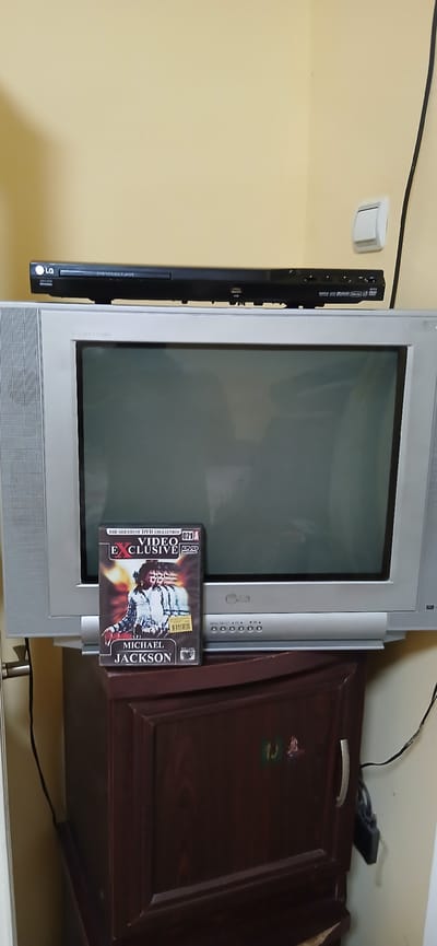 A DVD case featuring Michael Jackson is placed on the side of an old CRT television. The DVD is titled "The Greatest DVD Collection: Video Exclusive DVD" and prominently displays Michael Jackson's face. The television itself is silver and appears to be an older model, with a DVD player mounted vertically to its left side. The DVD player is black and has the LG logo. The scene is set against a plain, light yellow wall. To the right of the television, a dark wooden cabinet is partially visible, with a silver handle on its door. The overall impression is a somewhat cluttered, domestic setting, possibly a living room or bedroom, where entertainment devices are stored. The lighting suggests an indoor environment, though the time of day cannot be determined. No specific emotions are conveyed, and the image is a still life of these objects.