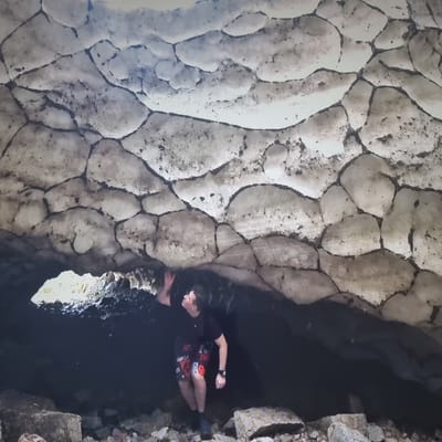 A man is standing inside a dark cave, looking up at the ceiling. The ceiling of the cave appears to be made of ice, with a honeycomb-like pattern of hexagonal shapes. The ice is light in color, with dark lines forming the boundaries between the shapes. The man is wearing a dark t-shirt and patterned shorts. He is reaching up with his right hand towards the ice ceiling. The cave floor is covered in rocks. The scene is dimly lit, with a small opening in the cave wall on the left side of the image. The location is not specified in the city of Aktobe, Kazakhstan.
