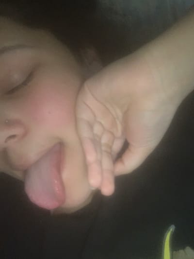 A close-up, angled image displays a person's partially visible face and left hand in low-light conditions. The individual's left eye is closed, and a silver stud piercing is visible on the left nostril. The tongue is extended from the mouth, exhibiting a pinkish-purple coloration. The left hand, with fingers lightly curled, rests against the left side of the face near the cheek and jawline. The visible skin is light-toned, with a reddish flush on the cheek. The background is dark and uniformly out of focus. A small, blurry yellow element is present in the lower right corner of the frame. The image provides no discernible environmental or architectural features to determine a specific location within the city of Lancaster, United States of America.