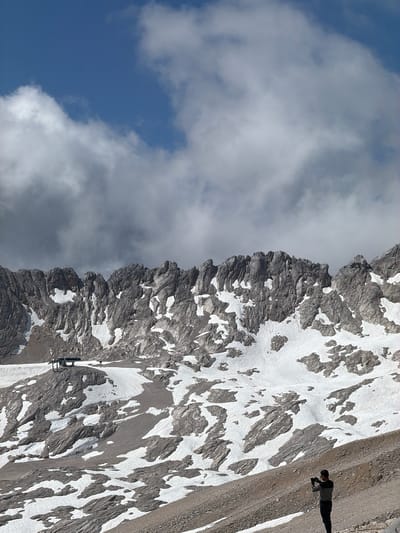 A lone individual stands on a gravelly slope in a mountainous, snowy landscape, holding a camera and appearing to take a photograph. The individual is wearing a striped long-sleeved shirt and dark pants. Above and behind the person are rocky mountain peaks with patches of snow. In the middle ground, a structure resembling a ski lift station is visible on a snow-covered plateau. The sky is partly cloudy with blue sky visible between white and gray clouds. The image appears to be taken in the vicinity of Wildermieming, Austria, based on the mountainous terrain and snow.