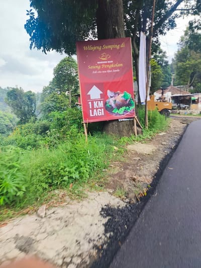 The image shows a sign on the side of a paved road. The sign is red and white with the words "Wilueng Sumping di Warung Nasi Saung Pengkolan" written in white on a red background. The sign also has a white arrow pointing up and the words "1 KM Lagi" written in white on a blue background. The sign is attached to a tree on the right side of the image. The road is paved and there is green vegetation on the left side of the image. There is a small building and a truck parked behind the sign. This scene is located near Lembang, Indonesia, at the coordinates -6.828 latitude and 107.607 longitude. The sign indicates a restaurant called "Warung Nasi Saung Pengkolan" is located 1 kilometer ahead. The image was likely taken by someone driving by on the road and the sign is designed to be visible to traffic.