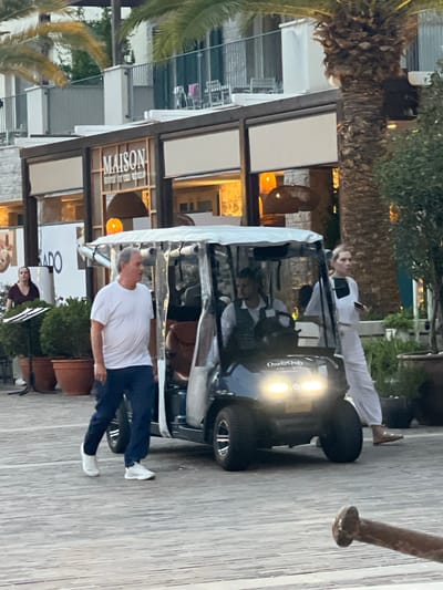 The image shows a man in a white t-shirt and dark pants walking on a wooden boardwalk. He is in the foreground, and a black golf cart is visible behind him. Two people are inside the golf cart; one appears to be a woman wearing white clothing and sunglasses, and the other is partially obscured. The golf cart has a light on in the front. Behind the golf cart is a building with a sign that says "MAISON". The building has large windows and appears to be a restaurant or shop. Palm trees are visible on the left side of the image. The lighting suggests it is either dusk or dawn, or a heavily overcast day. The scene appears to be a resort or a promenade area. The overall mood is casual and relaxed.