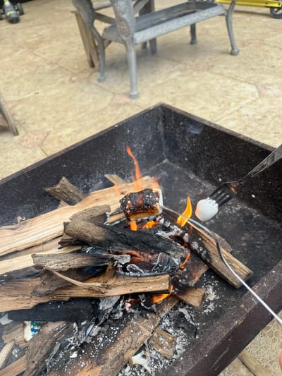 A fork holds a marshmallow over a small campfire in a metal fire pit. The fire pit is filled with burning logs and embers. The ground around the fire pit is paved with light-colored tiles. In the background, part of a metal bench and a yellow scooter are visible. The overall scene appears to be an outdoor setting, possibly a backyard or patio, in the city of Kaspiysk, Russia.