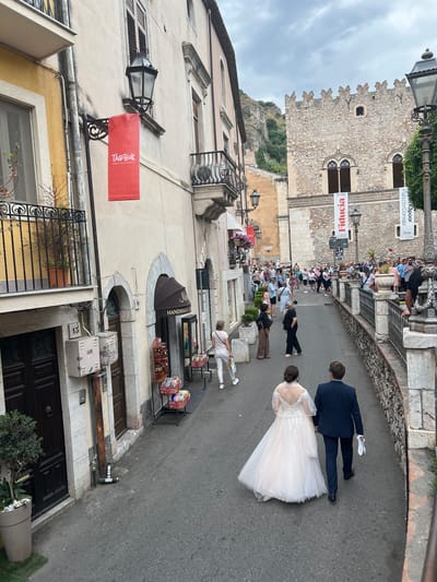 The image shows a view of a street in Taormina, Italy, likely a pedestrian area given the presence of many people walking. In the foreground, a bride in a white gown and a groom in a dark suit are walking away from the camera, hand in hand. The bride is holding a white cloth in her right hand. The street is paved with asphalt.

On the left side of the street, there are buildings with shops at ground level and balconies above. One building has a yellow facade. A red banner with "TAOBUK" written on it hangs from a lamppost. Another building has an awning for a shop labeled "M HANDMADE U..." with display cases selling colorful bracelets and other items. Above the shop, there are doors and windows, and a balcony with a metal railing.

On the right side of the street, there is a stone building with a fortified appearance, possibly a historical structure or palazzo. This building has arched windows and a crenellated top. Several banners hang from this building, including one with "FIDUCIA" and another with "PALAZZO CORVAIA." More people are walking and congregating in front of and around this building.

Further up the street, the landscape includes steep, green hills. The sky is overcast with clouds. Lampposts are visible along the street. The overall scene suggests a lively tourist area.