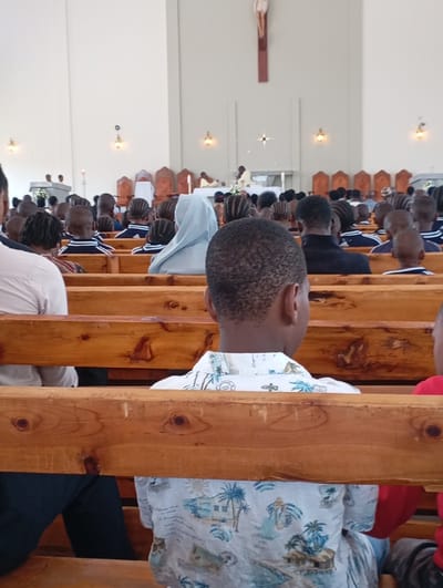 The image depicts a scene inside a church, likely located in Arusha, Tanzania, based on the provided coordinates. The image captures the back of a young man wearing a white shirt with a palm tree design, sitting on a wooden pew in the church. The church is filled with people, with the back of their heads and shoulders visible. The man is seated in the foreground, while other people are seated in the middle ground, and others are standing in the background.  The people in the background are engaged in a service, with one individual wearing a white robe standing at an altar. The ceiling is white, and the walls are also white.  The church is well lit, with several light fixtures visible. The image is taken at an angle, from the back of the man in the foreground, looking towards the front of the church.  There is a large cross on the wall behind the altar.  The man's focus appears to be on the service in the background.  The scene is calm and peaceful, depicting a typical religious service within the church.