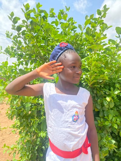 A young girl in a white uniform with a red sash and a blue cap poses outdoors. Her right hand is raised near her face. She is positioned in front of a lush green bush, with a blue sky and scattered clouds visible in the background. The ground appears to be dirt. This image was taken in Turu, Nigeria.