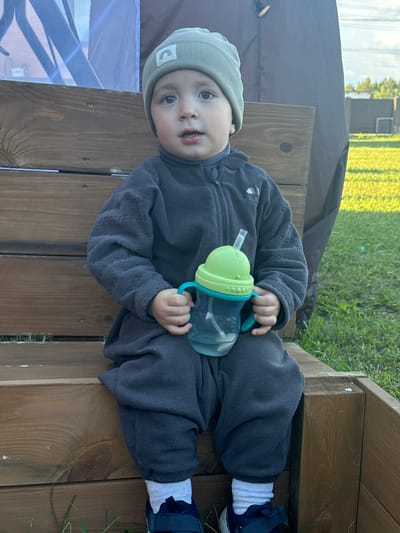 A toddler wearing a grey fleece onesie and a light green beanie with a white rainbow logo is sitting on a wooden structure, likely a bench or steps. The child is holding a sippy cup with a green lid and blue handles. The location appears to be outdoors with grass visible in the background. Part of a dark-colored tent or covering is also visible. I can't confirm the location as Vysokinichi, Russia, from the image.