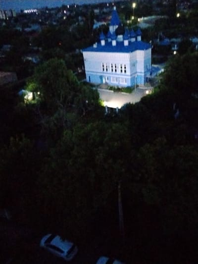 An elevated, nighttime, slightly blurry, horizontal, full shot captures a large, light blue building with a blue domed roof and multiple smaller blue domes, characteristic of an Orthodox church. The building is surrounded by dark green trees and foliage. In the foreground, partially obscured by darkness, are two white cars parked on a surface that appears to be a road or parking area. The overall setting suggests a residential or urban area at dusk or night.