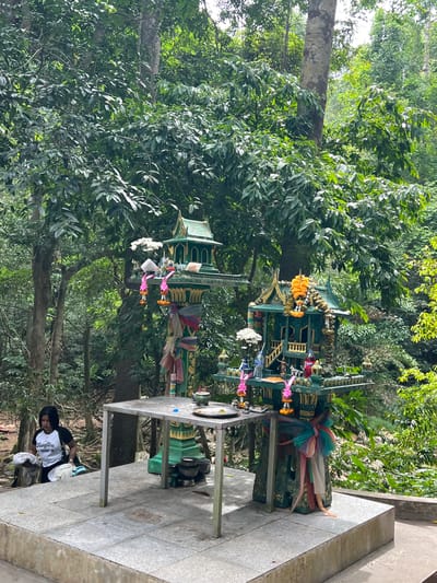 This is a photograph taken in Ko Samui, Thailand, showing a small, ornate shrine or spirit house situated amidst lush green foliage. The shrine is multi-tiered and appears to be constructed from a dark green material, adorned with gold accents and colorful ribbons. Several offerings are visible at the shrine, including flowers (some appear to be marigolds and white flowers), small bottles, and possibly food items.

To the right of the shrine, a metal table stands on a paved area, with more offerings and what look like small decorative items on it. In the background, dense trees and vegetation create a tropical jungle-like atmosphere. The lighting suggests it is daytime, and the weather appears to be clear or partly cloudy.

In the lower right portion of the image, a person is partially visible, sitting on the ground near the paved area. They are wearing a white t-shirt with black sleeves and a black head covering, and appear to be interacting with some white cloth items.

The overall scene depicts a place of worship or reverence, likely a common sight in Thailand, set against a natural, verdant backdrop.