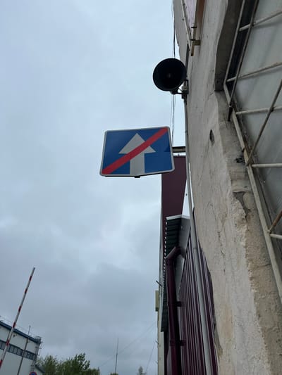 The image shows a "No Left Turn" road sign hanging beneath a concrete structure with a metal grate, likely an overhang of a building. A black loudspeaker is mounted nearby. The background includes a maroon-colored corrugated metal wall and a cloudy sky. In the lower-right corner, part of a white building and a red and white barrier are visible, suggesting a checkpoint or controlled access area. Based on the sign design, building styles, and other indicators, the location is likely Reutov, Russia. There are no people visible in the frame.