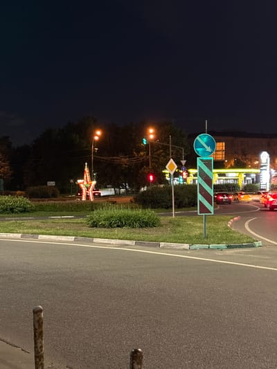 This is a nighttime, outdoor, street-level photograph taken in Mytishchi, Russia. The scene depicts a road with traffic and various signs and lights. A circular blue traffic sign with a white arrow pointing forward and slightly to the left is visible. Next to it is a rectangular sign with diagonal red and white stripes. Further down, a traffic light shows a red light. A large, illuminated star decoration is also present, likely a holiday display. The road is paved, and there is a grassy median with a curb separating it from the road. The background shows buildings and trees, illuminated by streetlights and building lights. The time of day is clearly night, with a dark sky. The weather appears clear, as indicated by the visibility and the presence of distant lights. There is no visible text that can be clearly deciphered in the image. The image appears to be taken from a slightly elevated, static perspective, suggesting the photographer was likely standing on a sidewalk or road shoulder.