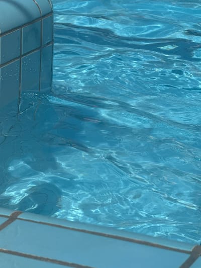 The image shows the interior of a swimming pool. The water is clear and blue, with refracted light patterns creating a shimmering effect. In the lower left, tiled pool walls are visible, while the edge of a pool ladder is visible on the right side. The location is presumed to be in Bat Yam, Israel, though there are no specific characteristics visible in the image to explicitly confirm this other than as requested. There are no people or actions taking place.