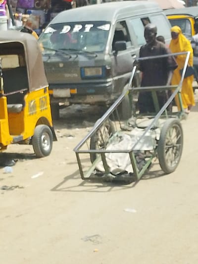 The scene depicts an outdoor street or market area in Fagge D1, Nigeria, during daylight hours. In the midground, a male subject in dark-colored attire is pushing a two-wheeled, metal-framed handcart containing light-colored material. A female subject, dressed in bright yellow headwear and clothing, walks behind him to the right. To the left foreground, the rear portion of a yellow three-wheeled auto-rickshaw with a brown fabric canopy is visible, bearing the text "Kano 0571" on its side. Behind the subjects, a dark grey Toyota minivan is present, featuring "DPT" in white lettering across its windshield. The ground consists of light-brown dirt or unpaved surface, with scattered litter. Indistinct structures and additional individuals are visible in the blurred background.