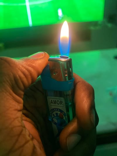 An extreme close-up shot shows a person's hand holding a lit lighter with a blue flame against a blurred green background that appears to be a television screen displaying a sporting event. The lighter has the brand name "AMOR" printed on it, along with a logo that includes the word "CHELSEA". The background's green hue and the presence of what looks like a stadium or field suggest the setting is likely indoors, possibly a living room or entertainment space, with a television as the primary light source and focal point of the background. The image is taken at night, likely in a residential area of Giginyu, Nigeria.
