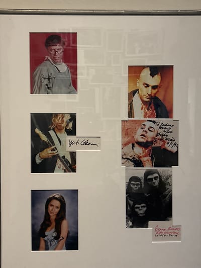 This image displays a collection of six autographed photographs, framed together under glass with a white mat. The frame itself is metallic. The setting is in the city of Venice, Italy.
The photographs are arranged in three rows:
**Top Row:**
* **Left:** A color photograph of actor Jerry Lewis portraying Professor Julius Kelp from *The Nutty Professor*. He is shown from the chest up, wearing glasses and a light-colored smock, against a red background. His autograph, "Jerry Lewis," is visible in the lower left portion of the image.
* **Right:** A color photograph of actor Robert De Niro as Travis Bickle from *Taxi Driver*, featuring his iconic mohawk haircut. He is depicted looking down, with visible bloodstains on his shirt.
**Middle Row:**
* **Left:** A color photograph of musician Kurt Cobain playing a guitar, with his long blonde hair partially obscuring his face. His autograph, "Kurt Cobain," is presented on a separate white slip of paper placed next to the photograph.
* **Right:** A color photograph of Robert De Niro again as Travis Bickle, covered in stage blood and looking upwards. An inscription on the photo reads, "To Richard Prince later Bobby De Niro 9/15/95."
**Bottom Row:**
* **Left:** A color portrait photograph of actress Jennifer Love Hewitt as a young woman, with long dark hair and wearing a white tank top, against a blue background. Her autograph, "Jennifer Love Hewitt," is visible on her top.
* **Right:** A black and white photograph depicting three individuals in ape costumes from *Planet of the Apes*. Two figures are prominent in the foreground, with a third slightly behind. An inscription on a separate white slip of paper below the photo lists names: "Pierre Boulle," "Kim Hunter," and "Roddy McDowall."