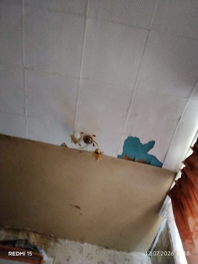 The image shows a damaged ceiling with peeling paint and visible water stains. The ceiling panels have a textured, geometric pattern. In the lower part of the image, a light fixture is attached to the ceiling, and the edge of a large, flat object, possibly a shelf or a piece of furniture, is visible. To the right, part of an orange curtain is seen. The bottom of the image contains a timestamp, "12.07.2026 11:48," and the text "REDMI 15." The scene appears to be indoors, likely in a residential setting, with evidence of disrepair.