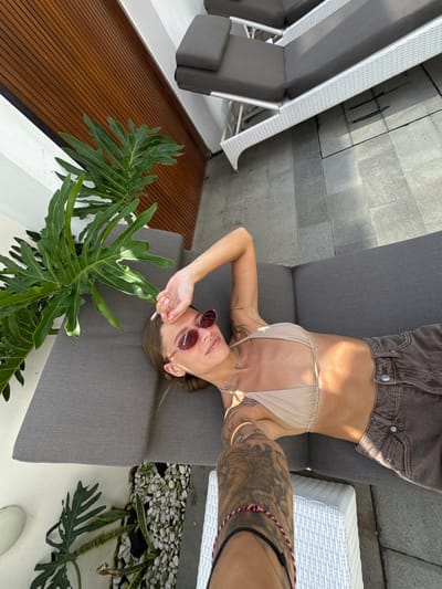 In Kediri, Indonesia, a woman lies on a gray outdoor sofa with a large green plant to her left. She wears a light-colored bikini top and dark shorts, with pink sunglasses on her face. Her right arm is raised, resting her hand on her forehead. Tattoos are visible on her arms and shoulder. The sofa is situated on a tiled patio, with a white woven side table to the right. In the background, a wooden-paneled wall and another gray sofa are visible. The scene appears to be an outdoor lounging area.
