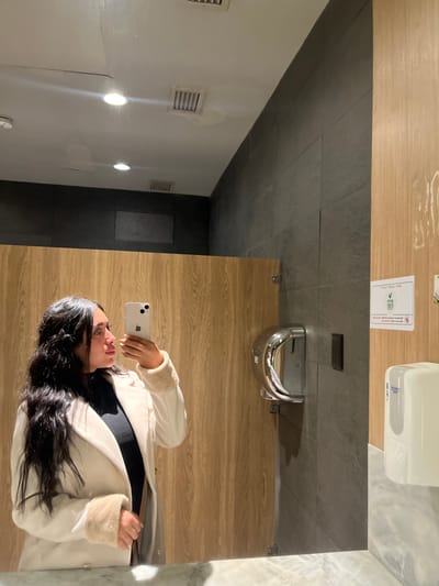 A woman is reflected in a bathroom mirror, holding a smartphone up to take a selfie. She's wearing a white coat with fur trim over a black top. The background shows a bathroom stall with light wooden panels, a black tiled wall, and a white hand dryer above the sink. The left side of the reflection reveals a white wall with vents and two recessed lights. The flooring appears to be light wood laminate.