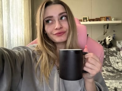A young Caucasian female, with long blonde hair and green eyes, is depicted from the chest up in an indoor setting in Reutov, Russia. She wears a light grey hooded zip-up sweatshirt and is seated in a pink gaming-style chair. Her right hand holds a black cylindrical mug, exposing pink-painted fingernails. Her facial expression includes slightly puckered lips and eyes looking upwards and to her left, indicating a contemplative or posed glance. Behind her, a sheer, light-colored curtain covers a window on the left. A white floating shelf on the wall to the right displays several framed photographs and a small white figurine. Below the shelf, a black and white panda plush toy rests near a grey and white patterned duvet or blanket. The wall is a light, neutral color. The image appears to be a selfie.
