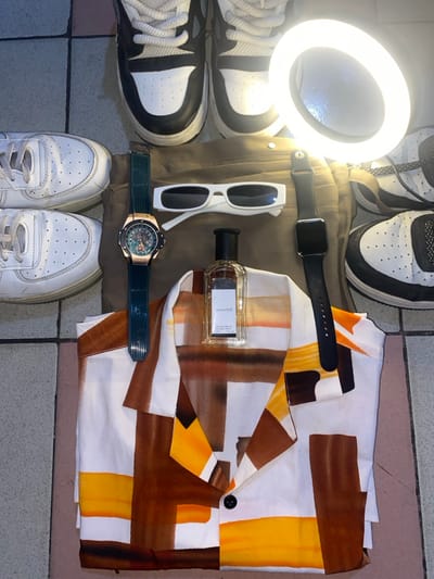 An overhead shot displays an organized arrangement of clothing and accessories on a light-colored tiled floor in Ngleshi Amanfro, Ghana. At the top of the frame, a pair of black and white high-top sneakers are positioned. To the left, two white low-top sneakers are stacked. A partially visible white and black low-top sneaker is situated to the right. Centrally, folded khaki-colored trousers serve as a base for other items. A folded white shirt featuring an abstract pattern of brown, orange, and yellow geometric shapes is laid over the lower portion of the trousers. A clear glass perfume bottle with a black cap, labeled "GALAXY 5B," stands upright on the shirt. To the left of the trousers, a gold-toned watch with a green textured band and an exposed mechanical movement is displayed. White rectangular sunglasses are placed horizontally across the upper trousers. To the right, a black smartwatch with a black band is positioned. A circular, illuminated ring light is present in the upper right quadrant, casting a bright glow over several items.