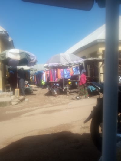 This image depicts an outdoor market scene in Serti, Nigeria, under a bright, clear blue sky, suggesting it is daytime with fair weather. The foreground is dominated by a dusty, unpaved ground with a large shadow cast from an unseen object, possibly a vehicle or structure.
In the mid-ground, several stalls are set up, covered by large umbrellas for shade. One stall prominently displays colorful fabrics and clothing items, hanging from a wooden structure. A person in a pink headscarf and patterned skirt is visible walking past this stall. Another stall to the left has a person in dark clothing standing behind a counter with more fabric displays. There are also bags and other merchandise visible on the ground near the stalls.
Further back, other market stalls and buildings with light-colored roofs are visible, extending into the background. The overall impression is of a bustling local market where goods are being sold under the open sky. The scene suggests a typical day in a Nigerian market, with vendors and customers engaged in commerce. There is no discernible text visible in the image.