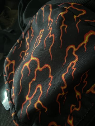 The image shows a close-up of a black fabric with a flame pattern in red and yellow. The fabric appears to be part of a mask or a piece of clothing. The flame pattern is abstract and stylized, with wavy lines and streaks of color. The texture of the fabric is visible, showing a woven material. The lighting is dim, casting shadows on the fabric and obscuring details in the background, which appears to be dark and possibly cluttered. There are no identifiable landmarks or elements that would suggest a specific location in Gelsenkirchen, Germany. The image is a static shot of an object.