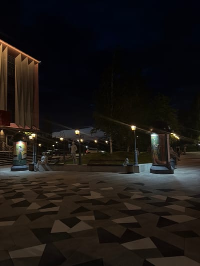The image depicts an outdoor public space at night, illuminated by streetlights. In the foreground, a patterned paved walkway with geometric shapes in varying shades of grey and black is visible. Several streetlights with warm yellow lamps are spaced along the walkway, casting light on the ground and surrounding elements.
To the left, a modern building with a facade featuring vertical ribbed elements and large windows is partially visible. Adjacent to the building is a set of stairs leading up. A large, illuminated cylindrical display case stands near the stairs, showcasing a poster with text and an image of a woman. Next to this display is a bench with a seated, life-sized statue of a person. Further down the walkway, more benches are present, some with statues of people sitting or standing.
In the background, trees with dark foliage form a canopy against the night sky. Cars are parked or driving on a road behind the benches and trees. The sky is a deep, dark blue with hints of clouds.
The scene appears to be a well-maintained urban area, possibly a park, plaza, or the entrance to a cultural institution. Based on the visual cues, including the style of architecture and public art, this location is identifiable as the square in front of the "Altynbash" Cinema in Naberezhnye Chelny, Russia. The statues are a notable feature of this public space.