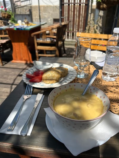 The image shows a table setting, likely in an outdoor area in Troyan, Bulgaria. The table is set with a bowl of soup, a plate with what appears to be fried cheese, tomato slices, and a small dipping sauce, a glass, a bottle of water, silverware wrapped in a white napkin, and a knitted placemat. The table appears to be made of dark wood. Behind the table, one can see a wooden chair and a glimpse of the surrounding environment, including buildings and greenery, indicating an outdoor restaurant or cafe setting in Troyan. The lighting suggests it is daytime. There are no people visible in the image.