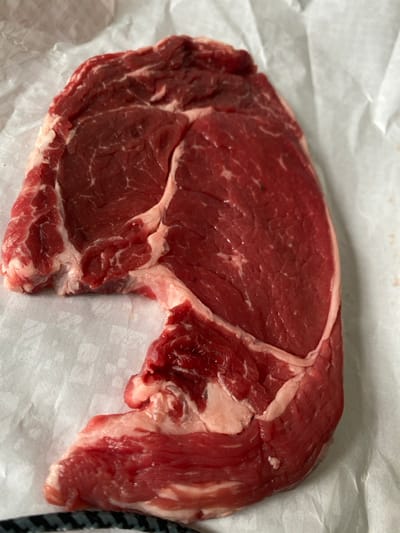 A single cut of raw red meat, likely beef steak, is depicted resting on a crumpled sheet of white butcher paper. The meat is predominantly deep red with irregular white marbling throughout and thicker bands of white fat along its irregular edges. One small, dark fleck is visible on the surface of the meat in the upper right quadrant. The paper exhibits creases and some discoloration suggestive of minor moisture absorption. No people are visible. No actions or interactions are occurring. The precise location within the city of Lille, France, cannot be determined from the visual evidence provided.
