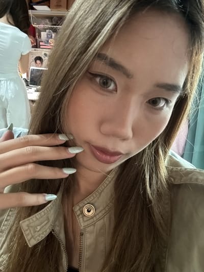 A young East Asian woman is taking a selfie. She has long, light brown hair, light brown eyes, and is wearing makeup. Her left hand is raised to her face, with light green fingernails visible. She is wearing a light brown collared jacket with a zipper and a button. In the background, a person in a white dress and a laptop are visible. The image appears to be taken indoors. The location is not discernable from the image.