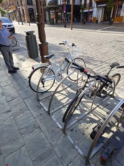 An outdoor street scene in Seville, Spain, shows a row of parked bicycles in a bike rack. In the foreground, a man in a light blue collared shirt and gray pants stands to the left, holding a mobile phone. Behind him, a white bicycle with a basket is parked in the rack, followed by a darker-colored bicycle. Further back, a dark gray trash receptacle stands near a lamppost. The ground is paved with cobblestones. In the background, buildings line the street, one of which features a McDonald's sign. People can be seen walking on the sidewalk. Sunlight casts shadows across the scene.