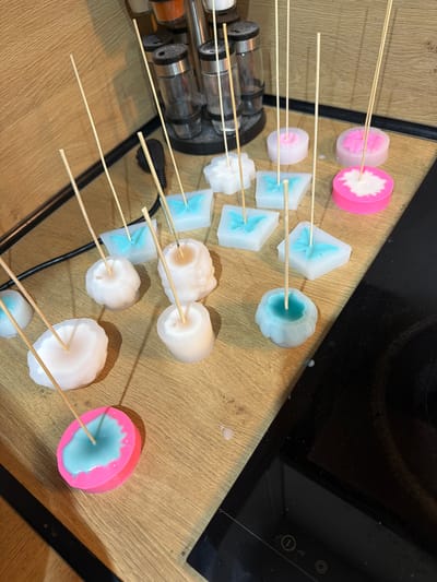 The image depicts an indoor scene on a light-colored wooden countertop, identified as being in Vladaya, Bulgaria. A collection of approximately 15 to 20 decorative wax items, likely candles or wax melts, is arranged on the surface. Each wax item has a slender, light-colored wooden stick inserted vertically into its center, functioning as a wick or a wick stabilizer during the setting process.
The wax items exhibit diverse shapes and color combinations. Several rectangular, translucent molds contain solidified light blue wax shaped like butterflies. Circular wax items are present, some featuring a solid white color, others with a vibrant pink exterior encompassing a white or light blue central core. Additionally, multiple irregularly shaped white wax items display textured or floral patterns, and at least one similar item is colored light blue. Small drips and solidified remnants of wax are visible on the wooden countertop, indicating recent pouring or handling activity.
In the upper background, on the same wooden surface, a multi-tiered metal spice rack holds several glass spice jars with dark lids. To the right of the wax items, a portion of a black induction hob is visible, displaying touch controls including a power button and circular heat indicators. The environment appears to be a kitchen or crafting space. No individuals are present in the image.