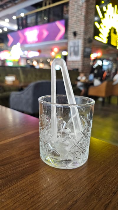 A close-up, eye-level shot features a clear glass tumbler filled with ice cubes and clear plastic tongs resting inside. The tumbler sits on a polished wooden surface, likely a table. The background is blurred, revealing an indoor setting with a brick wall on the right and a plush, dark-colored sofa on the left. The overall impression is of a bar or lounge environment. There is no indication of the location being Sevastopol, Ukraine.