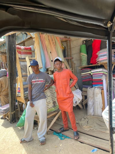 The image displays two young males standing within an outdoor market stall area in Zaria, Nigeria. The photograph is taken from a slightly elevated position, partially framed by a dark overhead structure, suggesting it was captured from inside a vehicle.
The male on the left wears a navy blue baseball cap, a grey t-shirt featuring "NEW ORLEANS FOOTBALL" text, light grey sweatpants, and olive green slide sandals. His hands are clasped together at waist level. The male on the right wears a white baseball cap with a red star emblem, an orange traditional long-sleeved tunic with matching trousers, and black slide sandals. His hands are placed on his hips. Both individuals face the camera with neutral expressions.
The background depicts a market stall structure constructed from wooden beams and concrete blocks. Various textiles are prominently displayed: multiple rolls of green, black, and red fabric are stacked vertically on shelves to the right. Below these, numerous piles of folded fabric in assorted colors and patterns are arranged. Overhead, several colorful and patterned fabrics are draped horizontally. A partially visible green and white commercial sign is situated behind the central figures. The ground consists of unpaved dirt, with loose wooden planks and a green plastic bag visible. Partial figures are present on the far left, dressed in light brown, and on the far right, wearing white, indicating additional individuals or activity within the scene.