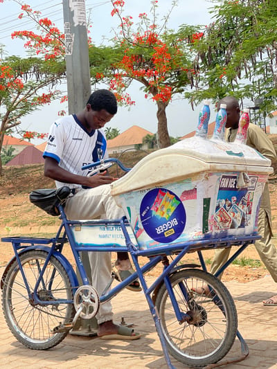 A man is sitting on a blue tricycle with an ice cream cooler in Kuchingoro, Nigeria. The cooler is white and covered in advertisements for Fan Milk products and the cooler has several yogurt drinks displayed on top. The man is wearing a white soccer jersey with blue trim and a black bag slung across his body. The tricycle is parked on a paved surface, and there is another man standing on the other side of the cooler. In the background, there are trees with red flowers and buildings.