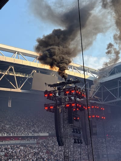 This media file depicts a large outdoor concert event, likely held in Dusseldorf, Germany. The scene is dominated by a massive stage structure adorned with numerous red lights and speakers, suggesting a professional musical performance. A large crowd of people is visible in the background, packed into the venue, indicating a popular event.

A significant amount of dark smoke is billowing from the top of the stage structure and spreading across the sky, obscuring parts of the blue sky above. This smoke is a prominent and dramatic element of the scene, suggesting a pyrotechnic effect, a malfunction, or possibly a controlled smoke release as part of the show.

The lighting from the stage, with its red lights, suggests the event is either taking place at dusk or under artificial lighting, creating a dynamic atmosphere. The presence of a large banner with visible German text adds a local touch. The text reads "IST EIN GROBARTIGER TAG!!" which translates to "IS A GREAT DAY!!". This cheerful message contrasts with the potentially dramatic smoke release. The overall impression is one of a high-energy event with a spectacular, possibly unexpected, visual element.
