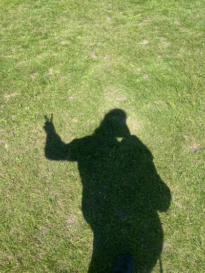 A shadow of a person is cast on green grass, with the person's left hand raised in a peace sign. The shadow shows the person is wearing a hat. The setting appears to be a park or open field. No specific location in Maidstone, United Kingdom can be determined from the image.