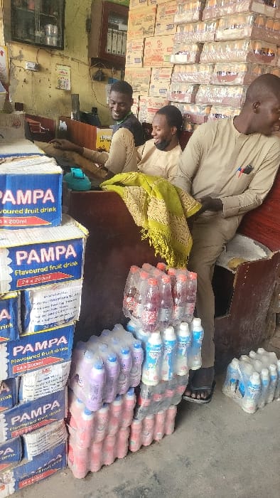 The image depicts an indoor retail space in Fagge A, Nigeria, characterized by stacked merchandise and three adult males interacting. On the left foreground, multiple blue and white cardboard boxes labeled "PAMPA Flavoured fruit drink, 24 x 200ml" are stacked vertically. To the right of these boxes, various plastic-wrapped bundles of bottled beverages are arranged on a concrete floor, including clear bottles with red contents, purple/pink bottles with blue caps, and smaller clear bottles with white caps.
Behind the beverage stacks, two men are seated or leaning behind a dark wooden counter. The man on the left wears a black short-sleeved shirt and light brown trousers, smiling and looking towards the right. The man in the middle wears a light brown traditional top and trousers, with a black face mask pulled below his chin, also smiling and looking right. A green patterned fabric with fringe is draped over the counter between the middle man and the man on the right.
The third man stands or leans on the right side of the counter, wearing a light brown traditional top with pens clipped to his chest pocket, dark trousers, and black flip-flops. He is smiling and looking towards the left, appearing to interact with the other two.
The background features a light-colored wall with some visible wear. On the left wall section, a dark framed item, an electrical outlet, and exposed wiring are present. To the right, a wooden cabinet with glass doors holds documents or files. Further right, the wall is extensively covered by stacks of cardboard boxes containing various products, suggesting a storage or sales area for bulk items. The overall lighting is consistent with an indoor setting during daylight hours.