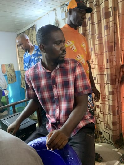 Within an indoor setting in Okene, Nigeria, three adult males are present.
In the foreground, an adult male with a dark complexion, short dark hair, and a trimmed beard and mustache is seated or crouched, facing slightly to his right. He wears a short-sleeved red, white, and blue plaid shirt and dark trousers. His hands rest on a blue plastic basin with embossed floral patterns.
Behind and to his right stands a second adult male with a dark complexion, wearing an orange baseball cap with a dark crown. He has on a short-sleeved orange t-shirt patterned with multiple yellow and white slipper-like designs. This individual is observed looking downwards and to his right.
Partially visible in the left background is a third adult male with a dark complexion and a bald or closely shaven head. He wears a short-sleeved blue button-up shirt featuring a white cityscape pattern and appears to be looking downwards.
The room features a white ceiling with exposed white electrical wiring and conduit runs. The wall on the left side is painted a light blue color, while a patterned brown curtain with a repeating geometric design covers the wall area to the right. A dark, rectangular electronic device is visible on a surface behind the central figure to his left, and a metal frame, possibly from a bed or bench, is also present.