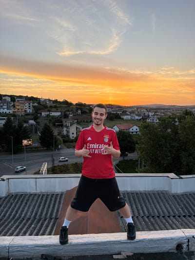 A young man stands on the corrugated metal roof of a building in Bragança, Portugal, against a sunset backdrop. He wears a red Benfica soccer jersey, black shorts, and white socks, and gives two thumbs up. In the background, a townscape with residential buildings stretches towards the horizon, with a road visible with cars and a sign in Portuguese. The sky shows a gradient from blue to orange at the horizon.
