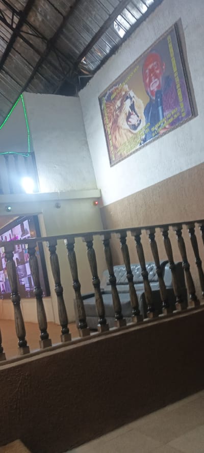 The image depicts an interior scene, likely in a building with a corrugated metal roof supported by metal beams. A framed picture hangs on a textured, off-white wall. The picture features a man with a microphone and a roaring lion. Text is visible on the picture, including "JUDAH IS THE LION OF THE TRIBE OF JUDAH GENESIS 49:9" and "THE RIGHTEOUS ARE BOLD AS A LION PROVERBS 28:1". A green light strip runs along a corner of the wall. In the foreground, a wooden banister with dark spindles lines a staircase. Below the banister, a large screen displays multiple video feeds, resembling a conference call or monitoring system. To the right of the screen, a portion of a tufted gray sofa is visible. The environment suggests a public space or an office setting. The location is identified as Rukuba Road, Nigeria.