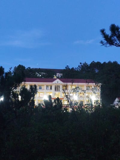 The image depicts a large, light-colored building with a red roof, illuminated by interior lights, as seen through the dark foliage of trees and bushes. The building appears to be multi-story with many windows, some of which are glowing with light. The architecture suggests it could be a school, a hospital, or a similar institutional building.
The scene is set at dusk or early evening, indicated by the deep blue of the sky and the artificial lighting of the building. The weather appears clear. The surrounding environment is densely vegetated, with dark green trees and bushes obscuring much of the lower parts of the building and framing the view. Pine trees are particularly prominent in the foreground and upper right portion of the image.
There are no people clearly visible in the frame. The activity taking place is not discernible, but the illuminated windows suggest occupancy or ongoing operations within the building.
The image was taken in Lạc Dương, Vietnam, which is a mountainous region known for its cool climate and pine forests. This context aligns with the visual cues in the photograph. No text is visible in the image. The overall mood is quiet and somewhat mysterious, with the illuminated building emerging from the shadows of the surrounding nature.
