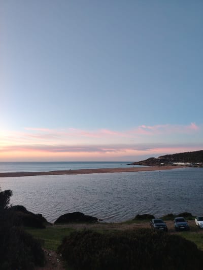 This image depicts a coastal landscape at dusk, specifically at Sidi Kacem Beach, near the mouth of Oued Tahaddart in Tanger, Morocco.
The sky occupies the upper two-thirds of the frame, transitioning from a light blue zenith to a horizon marked by horizontal bands of pink and orange stratocumulus clouds, characteristic of sunset. Below the sky, the vast expanse of the Atlantic Ocean is visible, meeting the horizon line. A long, narrow sandbar, light brown in color, separates the ocean from an inner, calm body of water, likely a lagoon or estuary. Two individuals are discernible as small silhouettes walking on this sandbar towards the left.
To the right, a rocky headland rises, covered in dark green vegetation. Scattered white buildings and illuminated structures are visible along the hillside, indicating human settlement or development. A winding road with visible lights ascends the hill. Additional small figures are present on the beach at the base of this headland.
In the foreground, a dark, elevated landmass covered in dense, low-lying dark green and brown shrubbery slopes downwards towards the inner body of water. Patches of lighter green grass are interspersed. Three vehicles are parked on a dirt patch near the water's edge on the right side of the foreground: two dark-colored SUVs/crossovers and one partially visible white car. The water in the lagoon exhibits minor ripples.