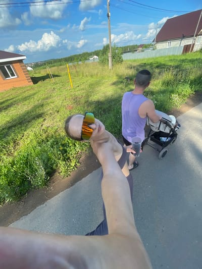 In a suburban setting in Podshivalovo, Russia, a person wearing sunglasses and a dark shirt is captured in a close-up, wide-angle selfie, with their arm extending towards the camera. Behind them, another individual wearing a sleeveless purple shirt pushes a baby stroller along an asphalt path. The path is bordered by a grassy area with wildflowers. In the background, houses with dark roofs and a light-colored metal fence are visible, along with a dense line of trees. The sky is blue with scattered white clouds, and power lines are present in the upper portion of the image.
