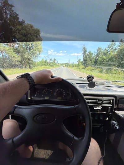 The image shows a first-person perspective from inside a car, with the driver's left hand on the steering wheel. The car is on a paved road that appears to be a highway, with trees and vegetation lining both sides. The sky is blue with some clouds. In the distance, other vehicles can be seen on the road. The dashboard of the car is visible, including the speedometer and other gauges, as well as a gear shift lever. The time on the dashboard is displayed as 14:30. The driver is wearing a watch on their left wrist. The scene is consistent with a person driving on a highway in a rural or suburban area. Based on the provided information, this scene is located on the road to the village of Garevaya, Russia.
