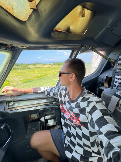 The image shows a person leaning out of the cockpit of what appears to be a small aircraft or helicopter. The person, a male with short brown hair, is wearing a black and white checkered long-sleeved shirt with red lettering that reads "Chec-Out". He is also wearing dark shorts and sunglasses.

The setting is the interior of a cockpit, with various control panels and instruments visible. The exterior view shows a bright, sunny day with a landscape of green fields, suggesting an outdoor location. The presence of a "Chec-Out" shirt could hint at the activity being a flight experience or tour. The overall lighting suggests it is daytime, likely mid-morning or afternoon, with clear weather. The location context provided, Blahbatuh, Indonesia, further places the scene in a tropical environment. The person's posture and outward gaze suggest curiosity or enjoyment of the view.