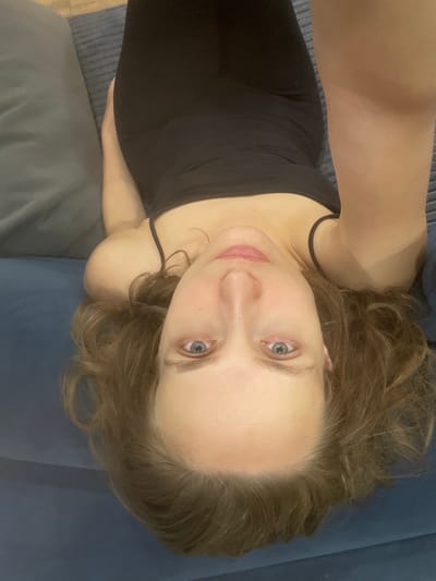 The media file shows a high-angle, close-up shot of a young woman lying on a couch in what appears to be an indoor setting. The woman has light skin, blue eyes, and medium-length, wavy brown hair that is spread around her head. She is wearing a black tank top with thin straps. Her expression is relaxed, with a slight smile. The couch is upholstered in a dark blue fabric, and a portion of a grey cushion or blanket is visible behind her. The lighting suggests it is likely daytime, as there are no visible artificial light sources contributing to the illumination. The image is likely taken in a home environment given the presence of the couch. No text is visible in the image. The location context provided is Подшивалово, Russia.