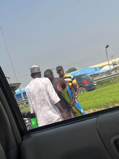 This image depicts an outdoor scene in Kagini, Nigeria. Three individuals are the primary subjects. One person, wearing a dark cap, a yellow and black striped shirt, and patterned shorts, is seated and appears to be holding a rolled-up mat or similar object. Another person, dressed in a purple patterned shirt, is standing behind the seated individual, also with a rolled object. A third person, wearing a white traditional outfit and a striped cap, is standing to the right, facing away from the camera.
The scene appears to be roadside, with a small black car visible in the background. Beyond the car, there is a grassy area and some structures, including what looks like a power line pole. The sky is a pale, hazy blue, suggesting a bright day, likely late morning or early afternoon. The overall impression is one of a casual, everyday moment. The expressions on the faces are not clearly discernible due to the angle and resolution, but the activity seems relaxed. A green object with some visible text, possibly a container, is partially visible on the right side of the frame, within a vehicle. The text visible on the green object appears to be "Wonderful" and a crescent moon with a star symbol. The image is taken from inside a vehicle, looking out through a window.